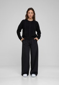 Zwarte kabelgebreide trui met ronde hals, gecombineerd met ruime zwarte broek en witte sneakers. Model staat met de handen in de zakken.