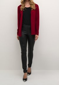 Rode cardigan over een zwart topje, gecombineerd met nauwsluitende zwarte leren broek en zwarte hoge hakken met metalen tips. Gladde texturen en een aangesloten ontwerp.