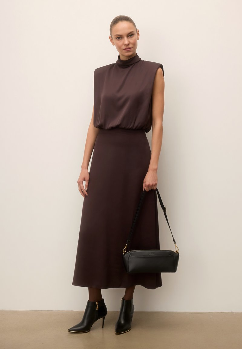 Femme portant une robe marron foncé sans manches tenant un sac à main noir, associée à des bottines noires, debout devant un mur clair uni.