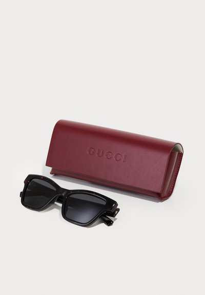 Sluneční brýle Gucci v černém želvovinovém designu, s tmavými skly, vedle maroon koženého pouzdra s embosovaným nápisem "GUCCI." Pouzdro je obdélníkové, hladké.