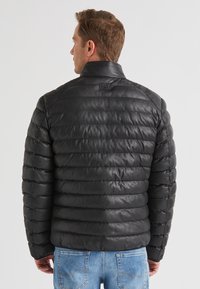 Schwarze gepolsterte Jacke mit Stehkragen, mit gestepptem Muster und einem Logosticker auf der Rückseite, kombiniert mit hellblauen Jeans.