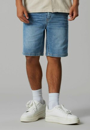 Man draagt lichtblauwe denim short, witte sportsokken en witte lage sneakers, staand op een effen grijze vloer en achtergrond.