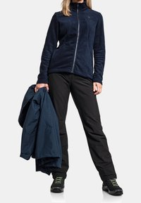 Marinblå fleecejacka med hög krage och dragkedja, matchad med svarta byxor. Håller en blå regnjacka. Har svarta skor med gröna detaljer.