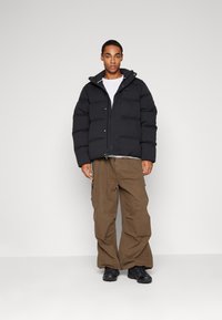 Jachetă neagră din puf matlasat, cu fermoar în față, guler înalt și un mic logo. Asortată cu pantaloni cargo relaxați maro și pantofi negri.