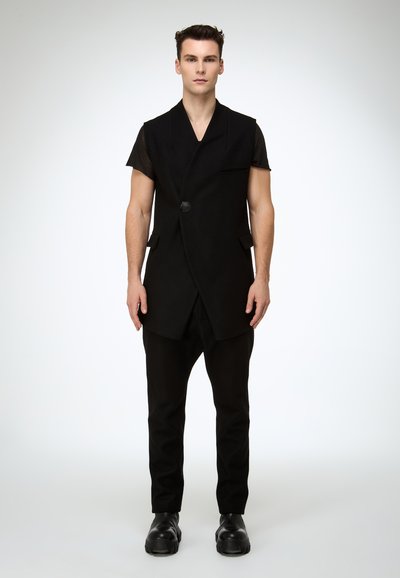 Schwarze ärmellose Weste mit V-Ausschnitt und seitlichen Taschen, kombiniert mit schmal zulaufenden schwarzen Hosen und schwarzen Schuhen, aus glattem, strukturiertem Stoff.