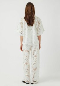 Set due pezzi bianco in pizzo con maniche ampie e pantaloni svasati con motivi floreali intagliati. Tessuto leggero e semitrasparente.