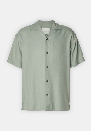 Chemise à manches courtes et col, en tissu vert clair, avec une patte de boutonnage à l'avant et une coupe décontractée. Portée avec un pantalon foncé et ample.