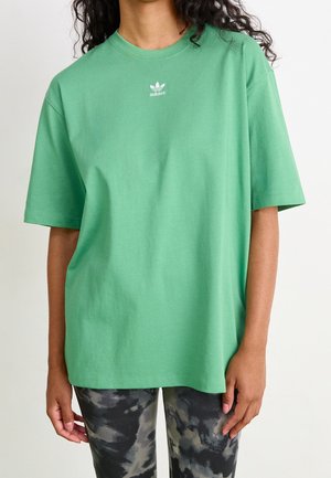 Person trägt ein locker sitzendes grünes Adidas-T-Shirt mit kurzen Ärmeln und schwarz-grauem Batik-Leggings, steht vor einem schlichten Hintergrund.