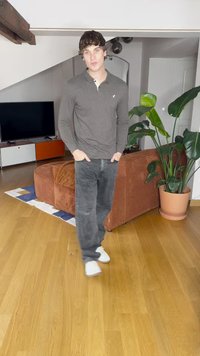 Graues langärmeliges Polohemd, dunkelgraue Hose und weiße Turnschuhe. Braunes strukturiertes Sofa und grüne Pflanze in einem Raum mit Holzboden.