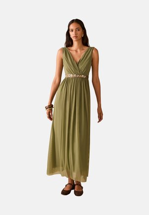 Donna che indossa un abito lungo verde oliva senza maniche con una fascia ricamata a fiori in vita, scarpe basse marroni e gioielli in oro, in piedi davanti a uno sfondo semplice.