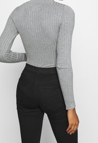 Personne portant un haut gris côtelé à manches longues ajusté, rentré dans un jean noir taille haute, debout avec une main sur la hanche.