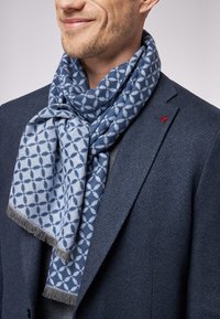 Lyseblå mønstret tørklæde med geometriske former og frynser, båret over en mørk blazer, med et synligt logo på jakkens revers.