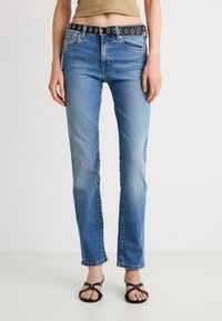 Niebieskie dżinsy denimowe o wąskim kroju, z czarnym paskiem ozdobionym srebrnymi okrągłymi elementami. Tkanina ma lekko wyblakły wygląd i minimalne przetarcia.