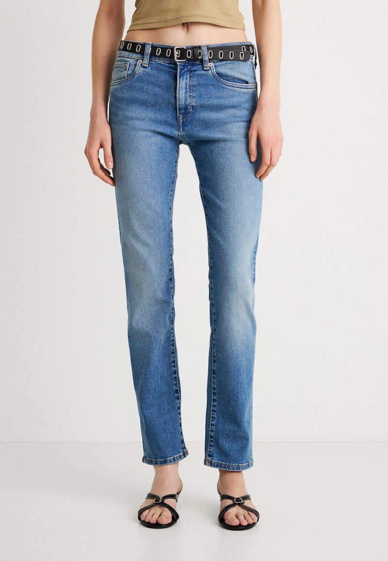 Pantalones vaqueros azules de corte ajustado, con un cinturón negro con detalles circulares plateados. La tela tiene un aspecto ligeramente desgastado y un mínimo desgaste.