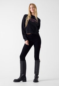 Czarna krótka bluza z logo, zestawiona z czarnymi obcisłymi jeansami i czarnymi skórzanymi kozakami za kolano. Gładka tekstura i dopasowany krój.