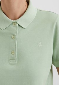 Vaaleanvihreä poolopaita, jossa on kolme nappia ja pieni brodeerattu "M OP" -logo vasemmassa rinnassa, päällä henkilö.