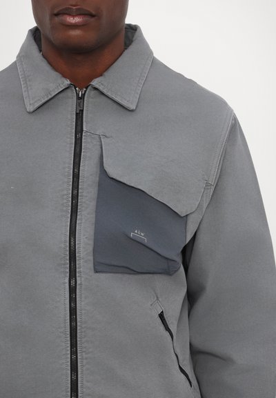 Veste zippée grise avec un col montant, dotée d'une grande poche avant en gris plus foncé et d'une poche latérale zippée. Détail de tissu texturé.
