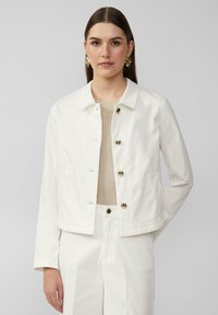 White denim jacket with a collared neckline, front buttons, and side pockets. Model wearing a beige top and matching trousers.