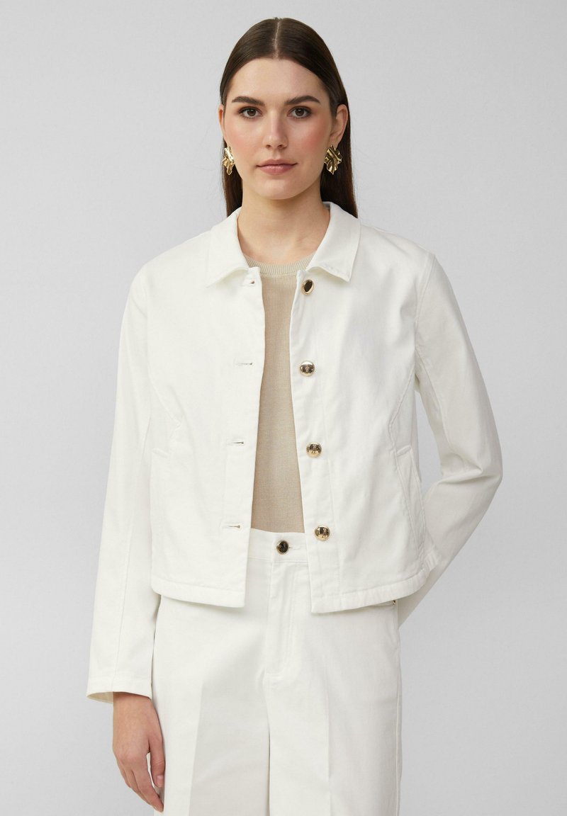 Weiße Jeansjacke mit einem Kragen, vorderen Knöpfen und Seitentaschen. Modell trägt ein beiges Oberteil und passende Hosen.