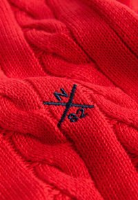 Punane kudumisest sviiter, millel on kaablitüübi muster ja tumesinisest lõngast broderi logo, näidates pehmet tekstuuri ja detailset käsitööd.