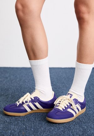 Jambes portant des chaussettes blanches montantes et des baskets Adidas Samba violettes avec des rayures blanches, debout sur un tapis bleu.