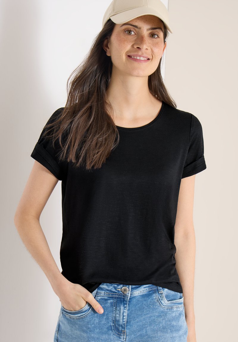 Jeune femme aux longs cheveux foncés portant une casquette beige, un t-shirt noir à manches courtes et un jean bleu, souriante avec une main dans la poche.
