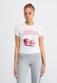 T-shirt blanc à manches courtes arborant le texte rouge "JORDAN" et des graphiques de cerises roses. Associé à un pantalon de sport gris avec un logo Nike.