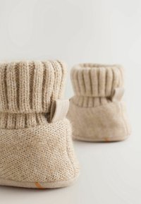 Chaussons pour bébé beige en tricot avec des poignets côtelés et des boucles en tissu, posés sur une surface claire unie.