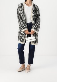 Person wearing black and white patterned cardigan, white top, dark jeans, beige ankle-strap shoes, holding small white handbag in front.