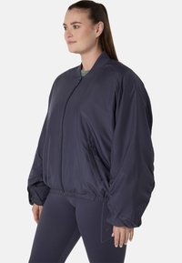 Frau in einer dunkelblauen oversized Bomberjacke mit Frontreißverschluss und Reißverschlusstaschen, kombiniert mit passenden Leggings, seitlich stehend.