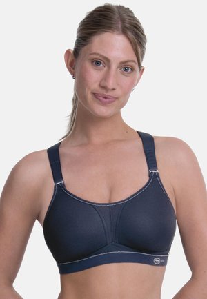 Ung kvinde med lyst hud og samlet hår iført en mørkeblå sports-bh med justerbare stropper mod en hvid baggrund.