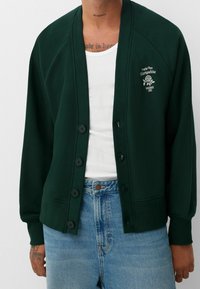 Cárdigan verde oscuro hecho de una tela suave, con cierre de botones en el frente, puños acanalados y un diseño bordado en la parte izquierda del pecho.