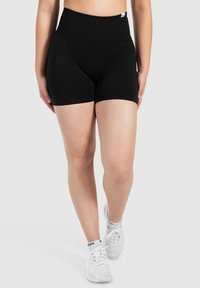 Pantaloni corti neri a vita alta realizzati in tessuto elasticizzato, con un design senza cuciture e una texture liscia. Abbinati a scarpe da ginnastica bianche.