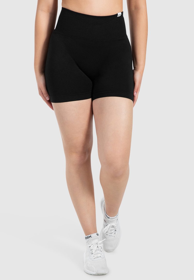 Pantaloni corti neri a vita alta realizzati in tessuto elasticizzato, con un design senza cuciture e una texture liscia. Abbinati a scarpe da ginnastica bianche.