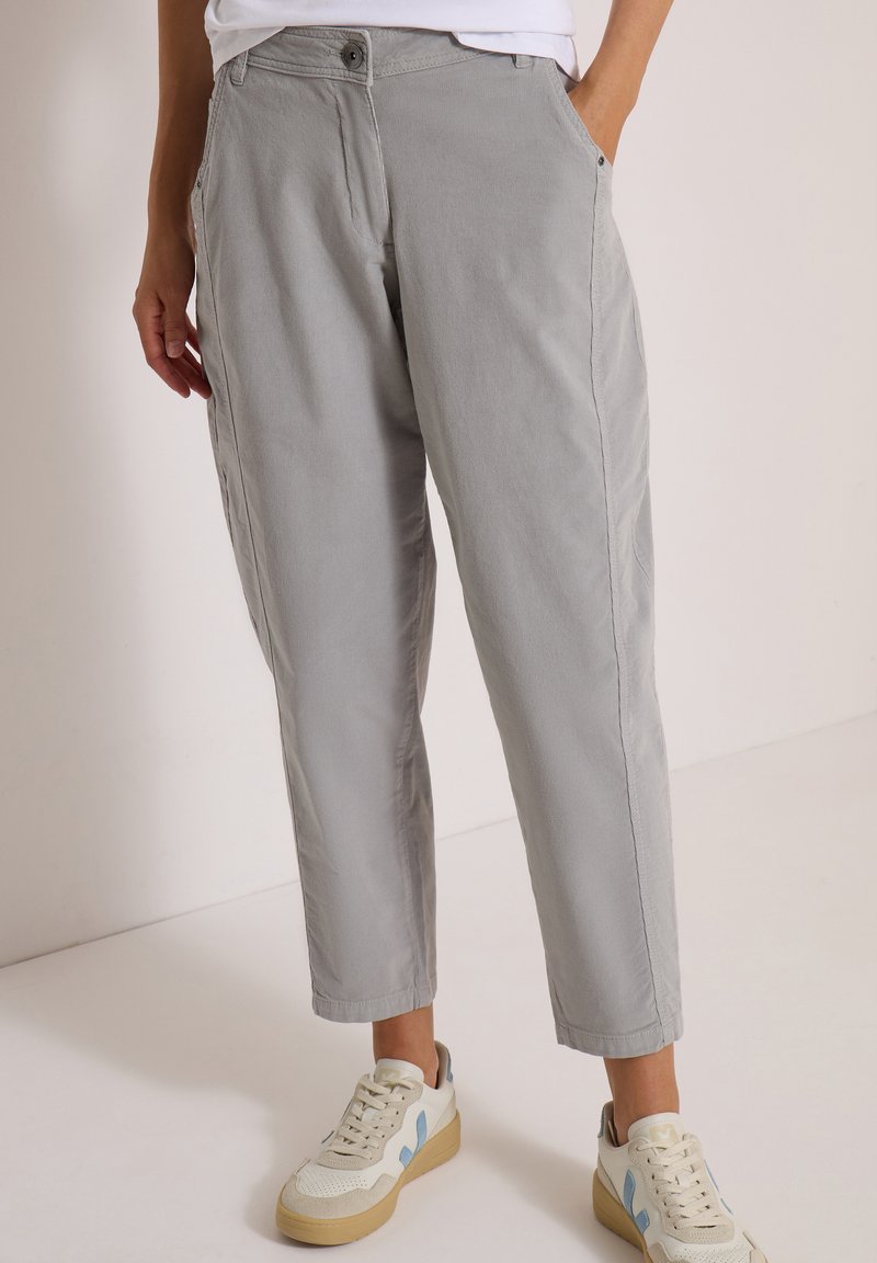 Personne portant un pantalon décontracté gris clair et des baskets blanches avec des accents bleus, debout avec une main dans la poche devant un fond uni.