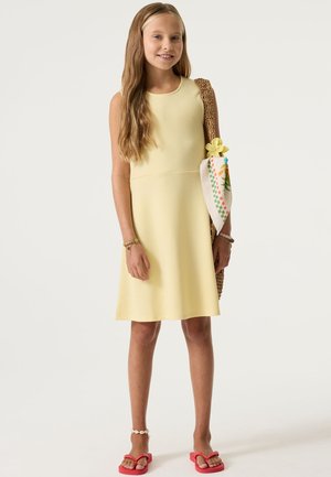 Vestido amarillo sin mangas con tela texturizada, silueta en línea A, bolso tejido a juego y chanclas rosas con pulsera blanca en el tobillo.