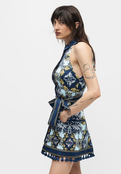 Άτομο με μακριά σκούρα μαλλιά και τατουάζ, φορώντας αμάνικο, διακοσμημένο μπλε και χρυσό φόρεμα με δεμένη ζώνη και φούντες στο τελείωμα, όρθιο πλαγίως.