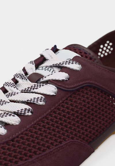 Κοντινό πλάνο ενός μπορντό αθλητικού παπουτσιού με κορδόνια σε λευκό και μαύρο σχέδιο και υφασμένο δίχτυ στην πλευρική επιφάνεια, πάνω σε λευκό φόντο.