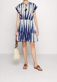 Woman wearing blue and white patterned short-sleeve dress, holding a cream woven tote bag, and brown flat sandals with open toes.