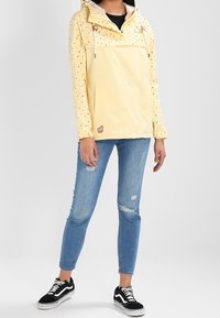 Gul lättviktsanorak med mönstrad övre del, dragskohuva, framficka, kombinerad med rivna blå jeans och svarta sneakers.