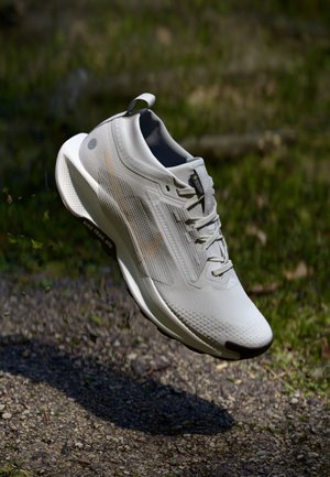 Basket de sport gris clair avec lacets en plein air au-dessus d'un chemin de gravier, montrant une semelle texturée et une tige en mesh respirant.