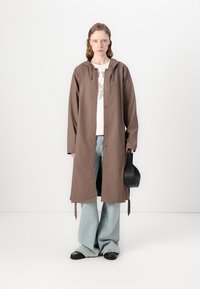 Brown waterproof long coat with a hood, button closure, and loose fit, paired with a white graphic t-shirt and light blue wide-leg jeans.