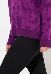 Pull en maille violet avec un motif texturé ton sur ton et des poignets côtelés, associé à un pantalon noir ajusté.