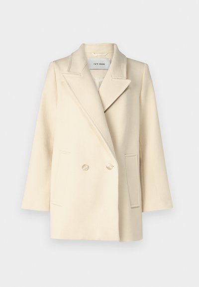 Cappotto doppio petto color crema con rever a punta, tasche laterali e bottoni chiari. Tessuto liscio e strutturato con texture minimale.