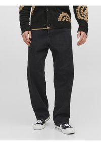 Hombre que lleva puestos unos vaqueros negros holgados, un cárdigan a rayas negro y marrón, y unas zapatillas en blanco y negro, de pie contra un fondo liso.