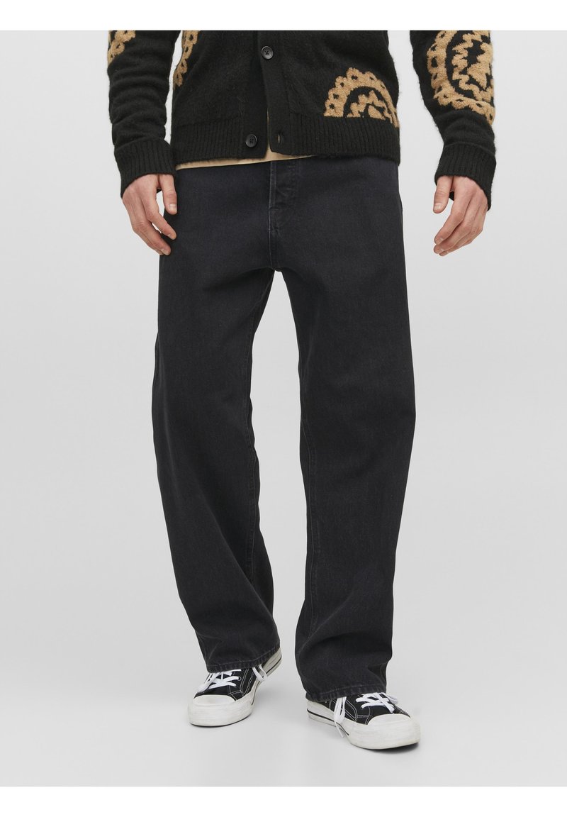 Hombre que lleva puestos unos vaqueros negros holgados, un cárdigan a rayas negro y marrón, y unas zapatillas en blanco y negro, de pie contra un fondo liso.