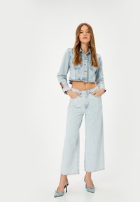 Chaqueta de mezclilla azul claro corta con cierres de botón y bolsillos en el pecho, combinada con pantalones de mezclilla azul claro de pierna ancha. Textura suave, diseño minimalista.
