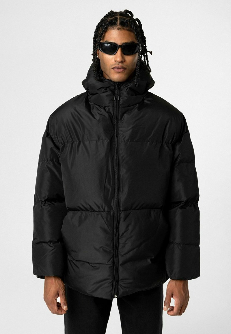 Doudoune noire avec un col montant, fermeture éclair à l'avant et manches larges. Texture lisse et brillante, rembourrée, offrant un look volumineux.