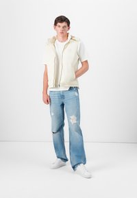 Helle, gefütterte Weste über einem weißen T-Shirt, kombiniert mit ausgebleichten blauen Jeans. Weiße Sneakers runden das Outfit ab. Einfaches, lässiges Design.