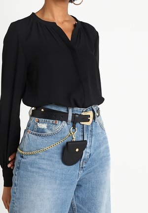 Femme portant un chemisier noir à manches longues rentré dans un jean bleu taille haute avec une ceinture noire et une petite pochette noire sur une chaîne dorée.